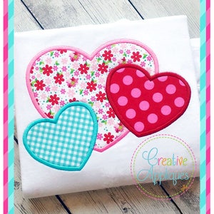 May include: Three felt hearts in different patterns, a pink heart with a floral pattern, a red heart with white polka dots, and a blue heart with a white checkered pattern. The hearts are on a white t-shirt.