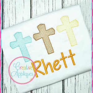 May include: A white t-shirt with three embroidered crosses in blue, brown, and yellow. The name "Rhett" is embroidered in orange below the crosses. The text "Creative Appliques" is embroidered in a circle below the name.