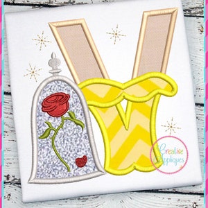 Könnte beinhalten: Stickerei mit einem gelben Buchstaben V mit Chevronmuster und einer Glaskuppel mit einer roten Rose darin. Das Design ist von kleinen goldenen Sternen umgeben.