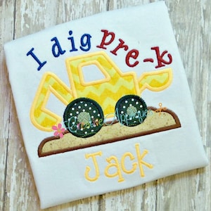 May include: A yellow and black embroidered design of a construction vehicle with the text "I dig pre-k" and the name "Jack" on a white t-shirt.