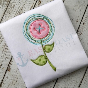May include: White fabric with an embroidered flower design. The flower features a pink button center with a blue cross, surrounded by teal and green stitching. The stem and leaves are green with polka dots.