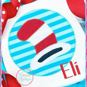 May include: A white t-shirt with a blue and white striped circle applique featuring a red and white striped hat. The name "Eli" is embroidered in red below the circle.