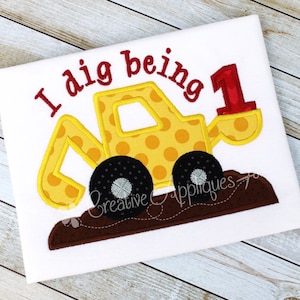 May include: Yellow and brown embroidered applique of a construction vehicle with the text "I dig being 1" on a white t-shirt.