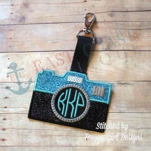 May include: A black and blue camera-shaped keychain with a silver glitter circle and the monogram "KKP" in turquoise. The keychain is attached to a black strap with a silver clip.