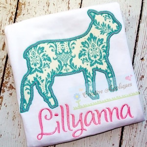 May include: A white t-shirt with a teal blue lamb silhouette embroidered on it. The lamb is filled with a white and teal blue damask pattern. The name "Lillyana" is embroidered in pink below the lamb.
