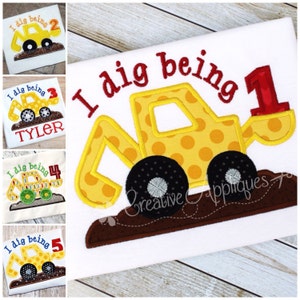 Op de afbeelding: Een geborduurde applicatie van een geel en zwart bouwvoertuig met de tekst "I dig being 1" op een wit T-shirt.