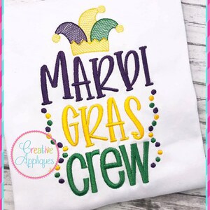 Op de afbeelding: Een wit T-shirt met een geborduurd ontwerp met een paarse en groene narrenmuts, de woorden "Mardi Gras Crew" in paars, geel en groen, en een rand van paarse en gele kralen.