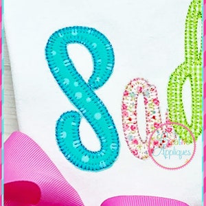 May include: A close-up of the letters "So" in a colorful applique style. The letter "S" is turquoise with white polka dots and the letter "o" is pink with a floral pattern. The letters are stitched onto a white fabric background.