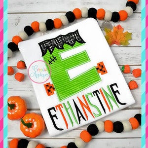 May include: A white t-shirt with a green Frankenstein-style letter "E" and the name "ETHANSTINE" embroidered in black. The shirt is surrounded by orange and black pumpkins and a garland of white and orange pom-poms.