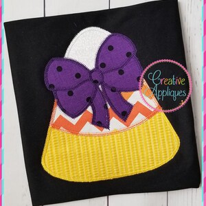 Op de afbeelding: Een paarse strik met witte stippen op een gele en oranje snoepmais applique. De applique is op een zwarte stoffen achtergrond.