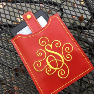 May include: Red leather e-reader case with a yellow monogram and a yellow strap. The case is on a black metal surface.