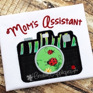 May include: Embroidered design of a camera with green and black stripes, a green lens with two red ladybugs, and the text "Mom's Assistant" in red.