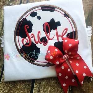 Può includere: Una maglietta bianca con un cerchio ricamato marrone e nero che presenta un motivo a stampa di mucca e il nome "Julie" in corsivo rosso. Un fiocco rosso con pois bianchi è attaccato alla maglietta.