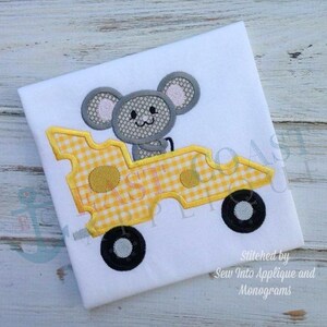 May include: A gray mouse driving a yellow and white checkered cheese car. The car has black wheels and the mouse is holding the steering wheel. The image is stitched on a white t-shirt.