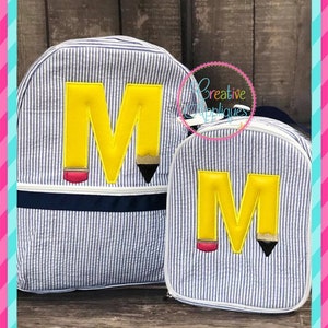 Könnte beinhalten: Ein blauer und weiß gestreifter Rucksack und eine Lunchbox mit einem gelben, bleistiftförmigen Buchstaben M auf jedem. Der Rucksack hat eine dunkelblaue Reißverschlusstasche und die Lunchbox hat einen weißen Reißverschluss.