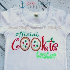 May include: White short-sleeved shirt with a red, green and brown embroidered design that says "official cookie taster" with two cookies on the left side of the text.