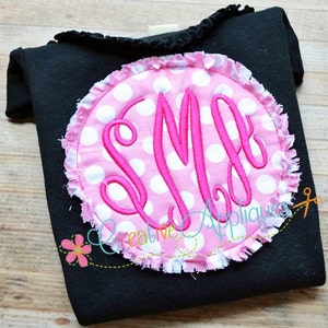 Op de afbeelding: Een roze en witte stippencirkel met een witte rafelige rand. De cirkel heeft een roze monogram "SM" geborduurd. De cirkel bevindt zich op een zwart shirt.