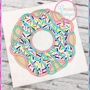 Könnte beinhalten: Ein rosafarbener und weißer Donut mit blauem Zuckerguss und bunten Streuseln. Der Donut ist auf einem weißen Stoff gestickt.
