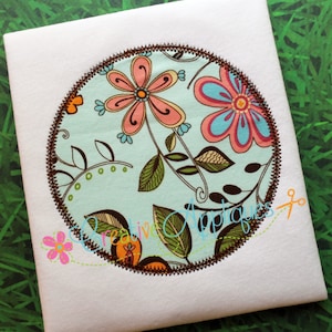 May include: A round embroidery design featuring a floral pattern with pink, blue, green, and brown flowers on a light blue background. The design is surrounded by a brown stitched border.