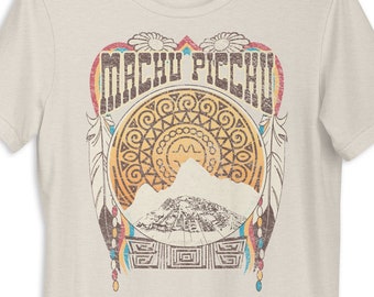 May include: A beige t-shirt with a vintage-style graphic of Machu Picchu, featuring a mountain range, a sun, and a circular design with geometric patterns. The text "Machu Picchu" is written in a stylized font.