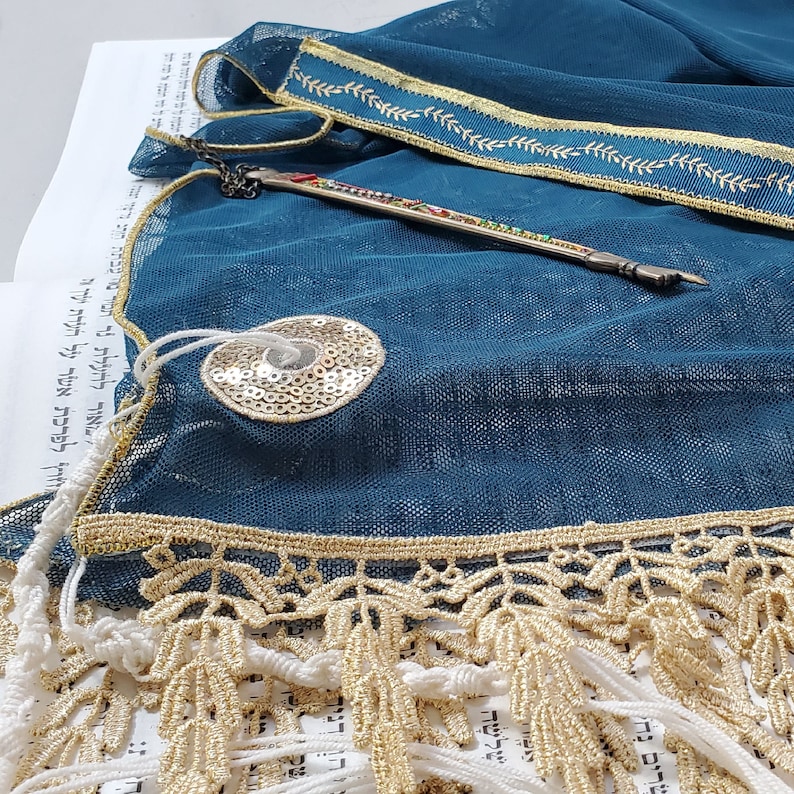 May include: A teal blue prayer shawl with gold trim and fringe. The shawl is laid out on a white surface with Hebrew text. A silver pointer with colorful beads is also visible.