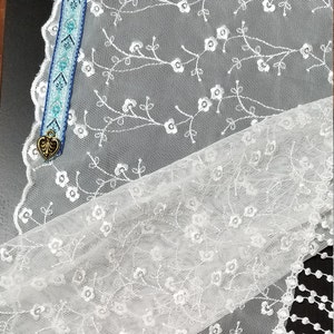 May include: White lace fabric with floral embroidery and a decorative edge. A blue and white patterned bookmark with a gold heart charm is visible. The lace has a delicate, airy appearance, suitable for crafting or decor.