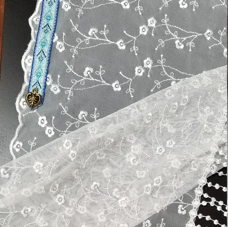 May include: White lace fabric with floral embroidery and a decorative edge. A blue and white patterned bookmark with a gold heart charm is visible. The lace has a delicate, airy appearance, suitable for crafting or decor.