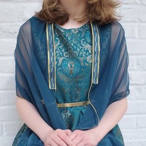 May include: A teal blue sheer shawl with gold trim over a teal blue and gold brocade dress. The dress has a gold belt.