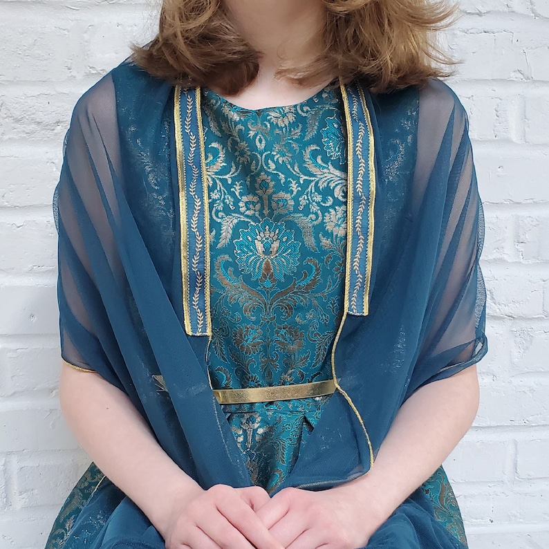 May include: A teal blue sheer shawl with gold trim over a teal blue and gold brocade dress. The dress has a gold belt.