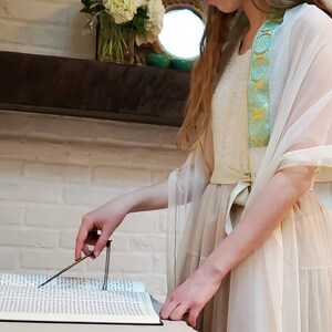 May include: A woman wearing a white dress with a sheer overlay and a blue and yellow sash is holding a pointer over an open book. The book is filled with Hebrew text.
