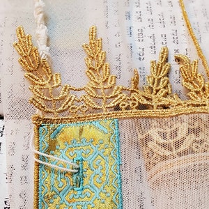 May include: A white fabric with gold embroidery and a blue and gold patterned fabric square. The fabric has a delicate, lacy texture.