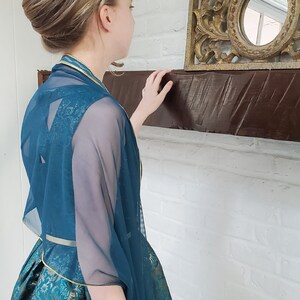 May include: A teal blue sheer shawl with gold trim. The shawl is draped over a person's shoulders and is worn over a gold and teal patterned dress.