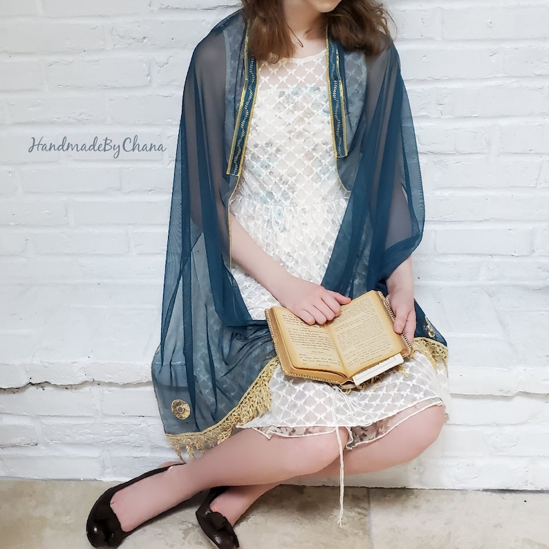 May include: A teal blue sheer shawl with gold trim and fringe, worn over a white lace dress. The person is holding a book.