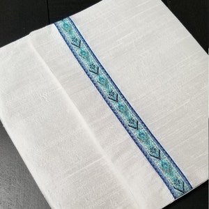May include: White rectangular textile with a decorative blue and teal patterned stripe running diagonally across the center. The stripe features geometric designs and is bordered by a darker blue trim. The textile appears to be folded.