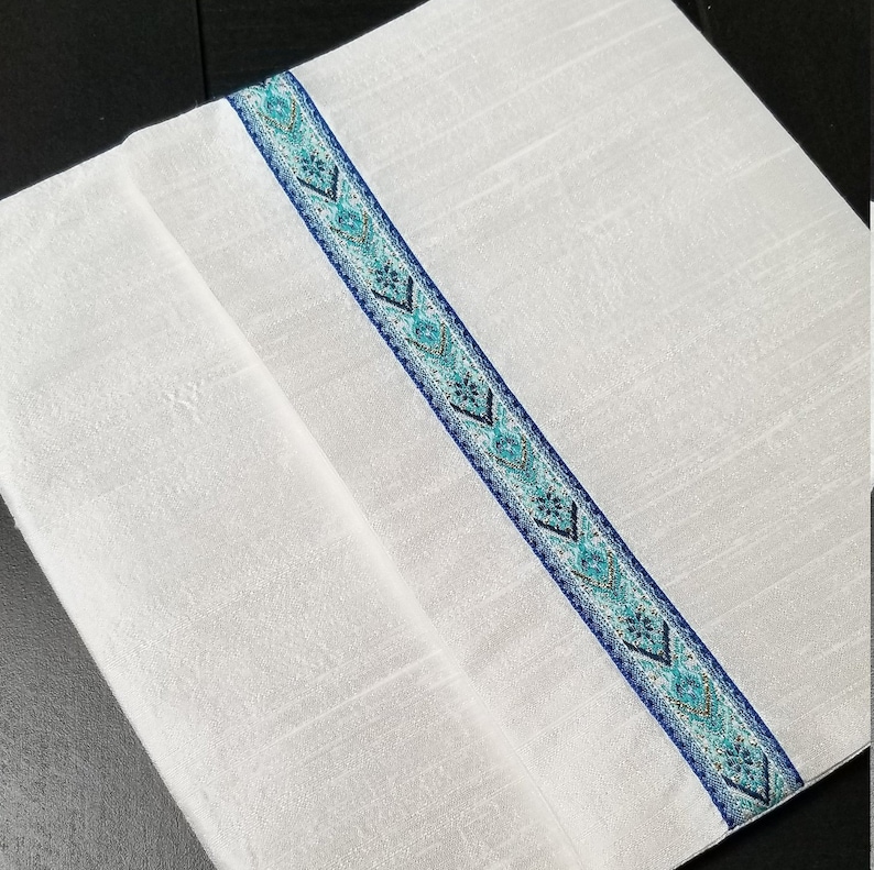 May include: White rectangular textile with a decorative blue and teal patterned stripe running diagonally across the center. The stripe features geometric designs and is bordered by a darker blue trim. The textile appears to be folded.
