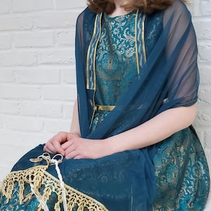 May include: A teal blue dress with a gold belt and a matching teal blue sheer shawl. The dress has a floral pattern and gold trim.