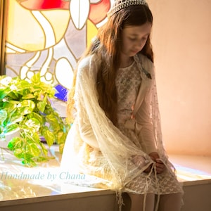 May include: A young person wearing a cream-colored dress and a delicate lace shawl, adorned with a crocheted crown. The image is set against a backdrop of a stained-glass window and includes the text "Handmade by Chana".