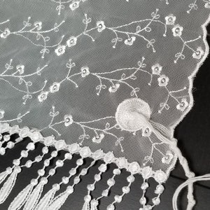 May include: Close-up of white lace fabric with floral embroidery and a scalloped edge. The delicate design features embroidered flowers and leaves, with a fringe of tassels along the bottom. A circular embroidered detail is also visible.