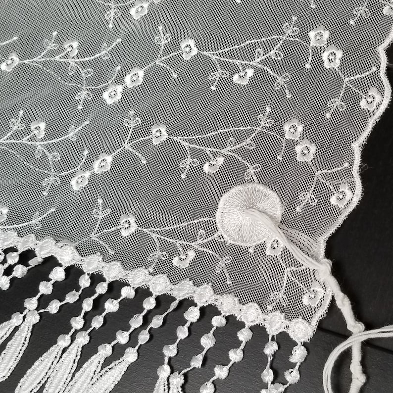 May include: Close-up of white lace fabric with floral embroidery and a scalloped edge. The delicate design features embroidered flowers and leaves, with a fringe of tassels along the bottom. A circular embroidered detail is also visible.