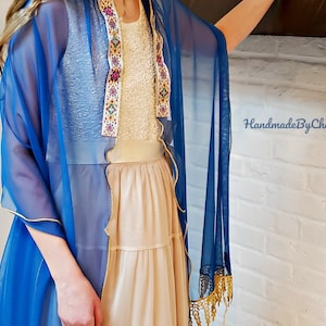 May include: A beige layered dress with a blue sheer shawl trimmed with gold. The dress has a beaded bodice and a tiered skirt. The shawl drapes over the shoulders and has gold fringe. The text "HandmadeByChana" is visible.