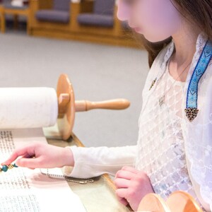 May include: A person is reading a scroll with a wooden handle. A blue and gold decorative ribbon is draped over the person's shoulder. The person is wearing a white lace top and a silver necklace.