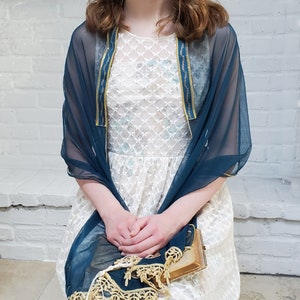 May include: A teal blue sheer shawl with gold trim and a white lace dress underneath. The shawl is draped over the shoulders and the dress is visible through the fabric.