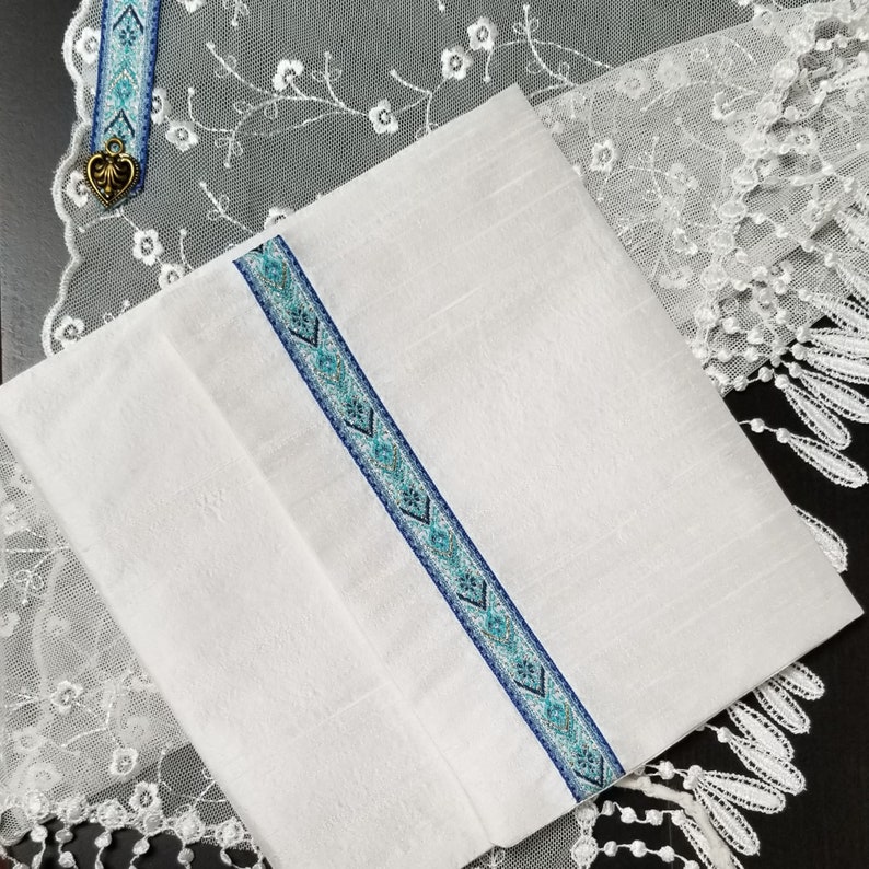 May include: White linen fabric with a decorative blue and teal patterned ribbon. A matching bookmark with a heart charm is visible in the upper left corner. The fabric is folded and placed on a white lace surface.