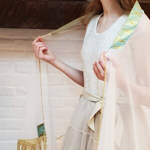 May include: A sheer white shawl with a gold and teal trim. The shawl is draped over a person's shoulders and is tied at the waist with a gold ribbon.
