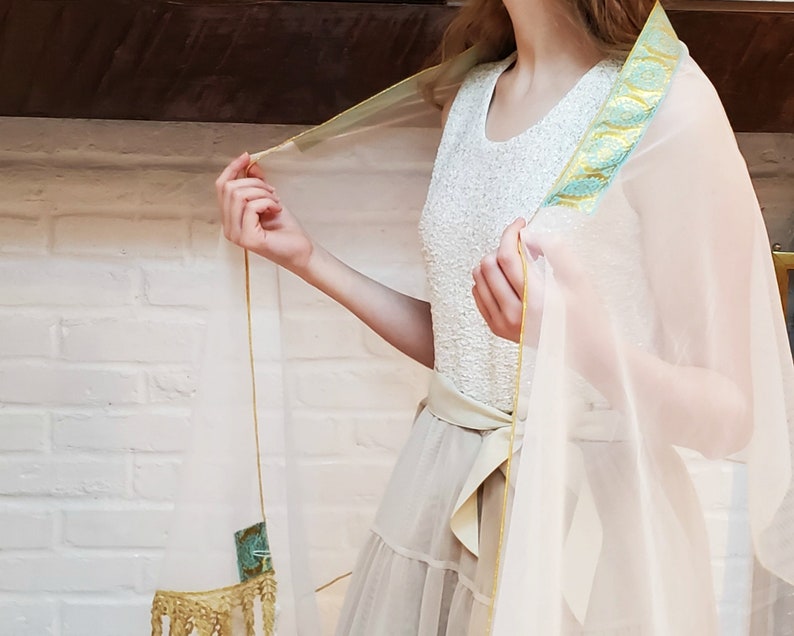 May include: A sheer white shawl with a gold and teal trim. The shawl is draped over a person's shoulders and is tied at the waist with a gold ribbon.