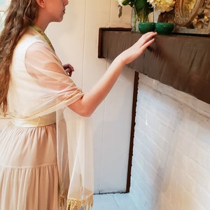 May include: A woman wearing a beige dress with a white sheer shawl draped over her shoulders. The shawl has a gold trim and fringe. She is standing in front of a white brick fireplace with a wooden mantel.