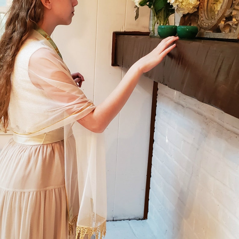 May include: A woman wearing a beige dress with a white sheer shawl draped over her shoulders. The shawl has a gold trim and fringe. She is standing in front of a white brick fireplace with a wooden mantel.