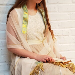 May include: A woman wearing a white dress with a sheer beige shawl and a green and gold trim. She is holding a book in her hands.