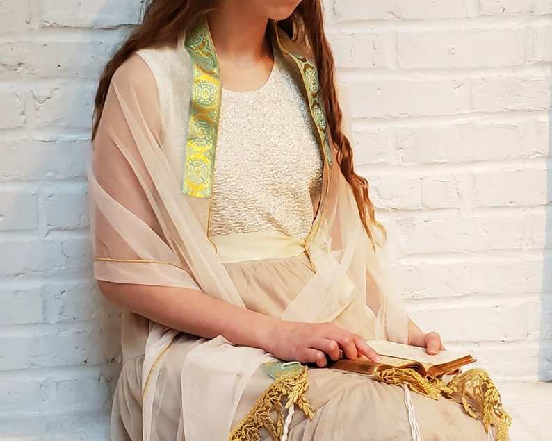 May include: A woman wearing a white dress with a sheer beige shawl and a green and gold trim. She is holding a book in her hands.