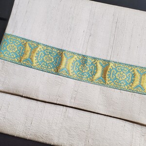 May include: A white fabric envelope with a decorative gold and teal trim. The trim features a repeating geometric pattern.